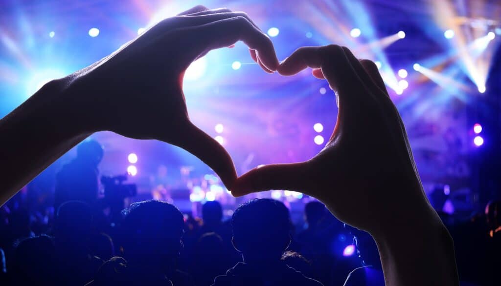Hands forming a heart shape with a concert and stage lighting in the background.