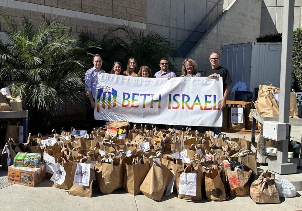 A group of congregants hold a Beth Israel banner in front of a large pile of assembled food donation bags.