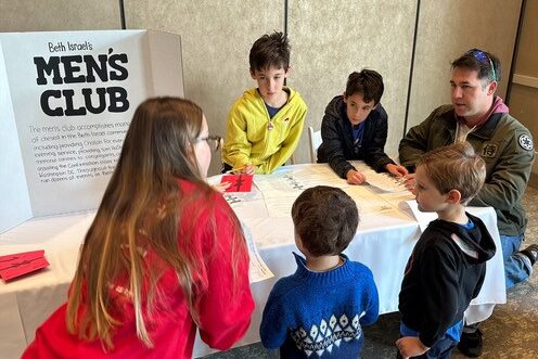 A group of children gather around an information table about Men's Club at Beth Israel.