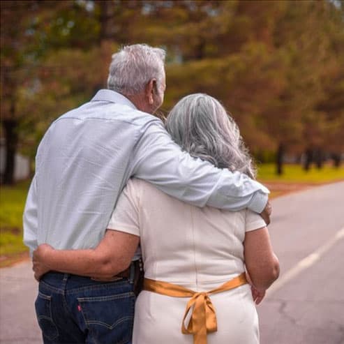 Older couple with arms around each other, walking on a tree-lined road.