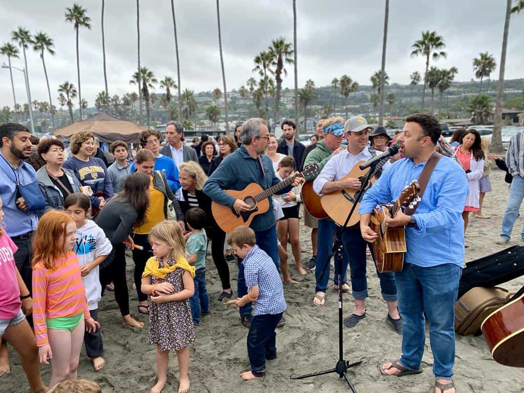 Kids dance while adults sing and play guitar on the beach during a Tashlich celebration.