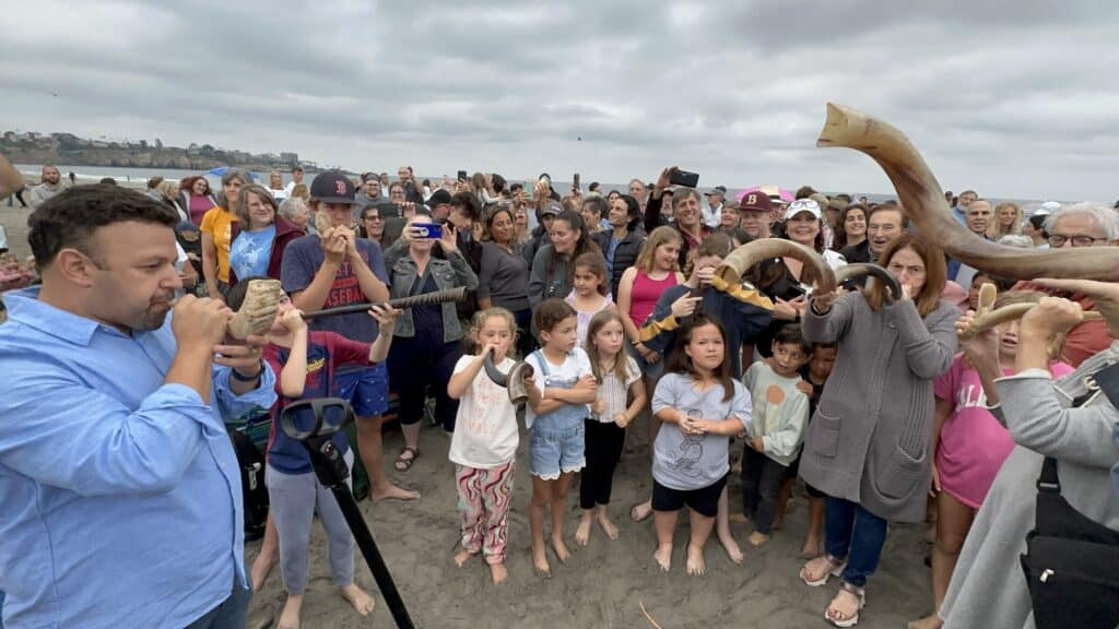 A group of congregants, along with a Rabbi, blow shofars during a Tashlich service on the beach.