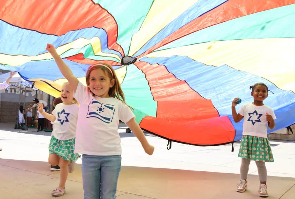 Three preschool students play under a parachute wearing shirts that celebrate Israel's Independence Day.