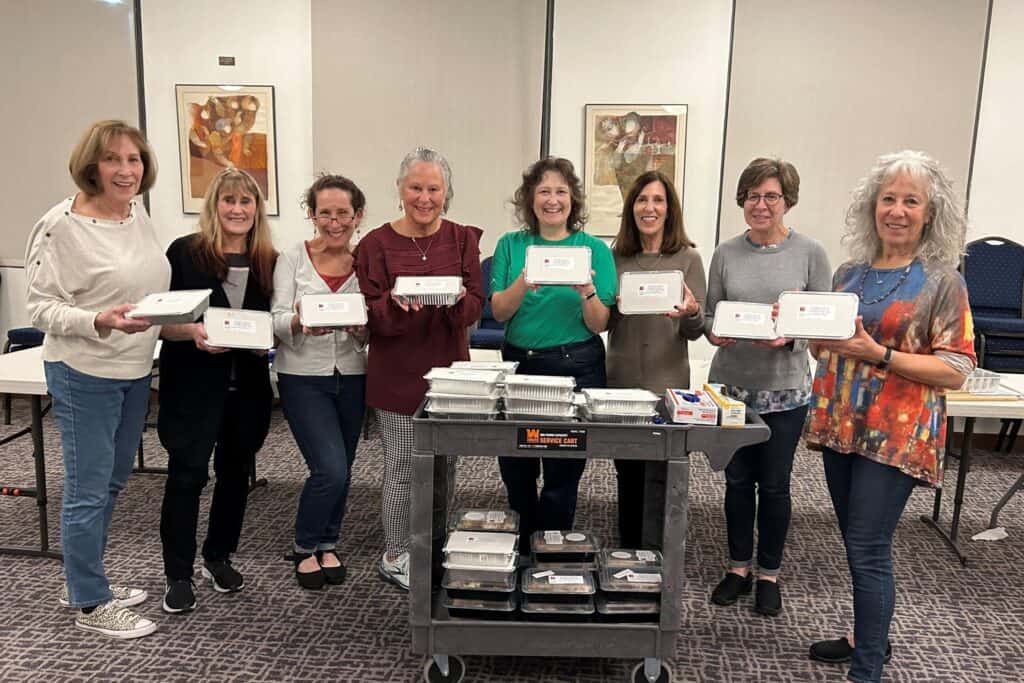 A group of women stand together holding the meals they have made for the Fill The Freezer program.