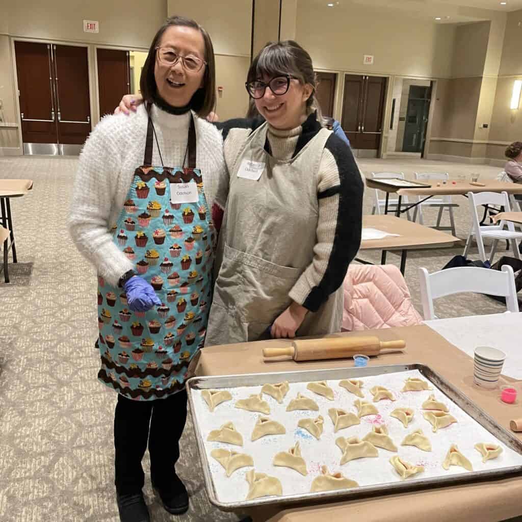 Two women stand next to a sheet of hamentashen they have made.