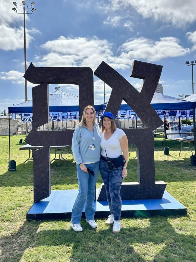 Two women pose in front of a sculpture made out of Hebrew letters.