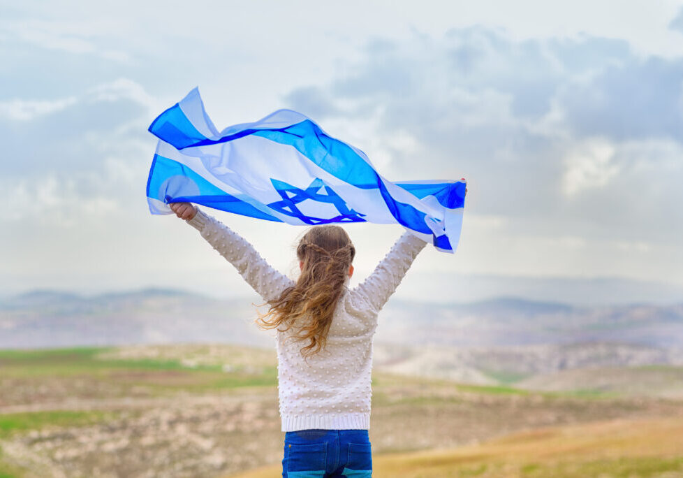 Girl standing in open field, with the flag of Israel held over her head.