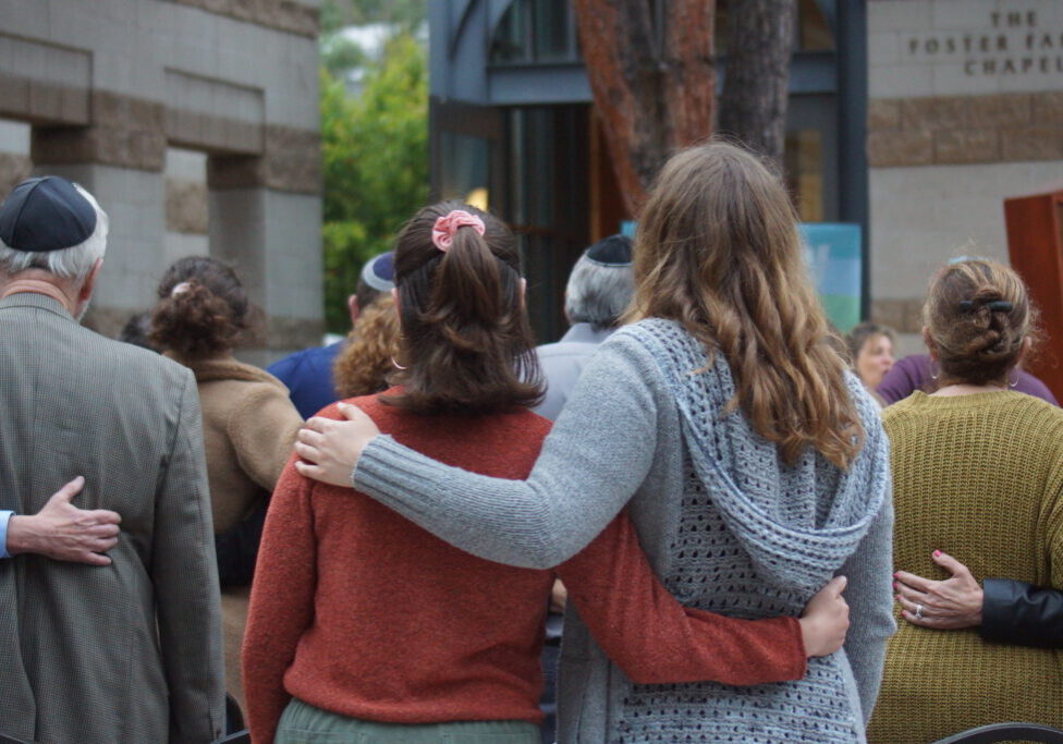 Two women stand arm in arm, their backs facing the camera.
