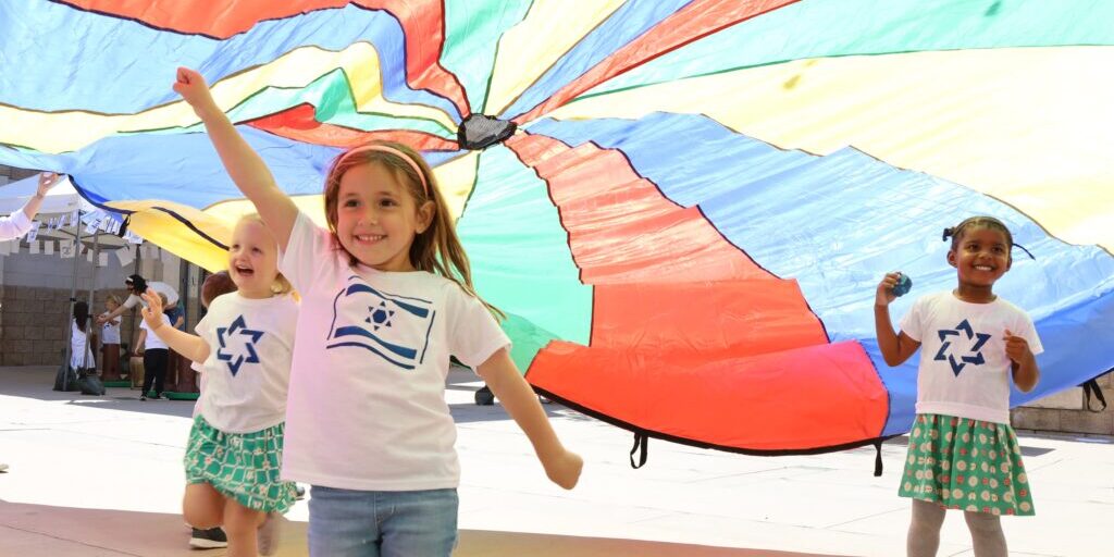 Three preschool students play under a parachute wearing shirts that celebrate Israel's Independence Day.